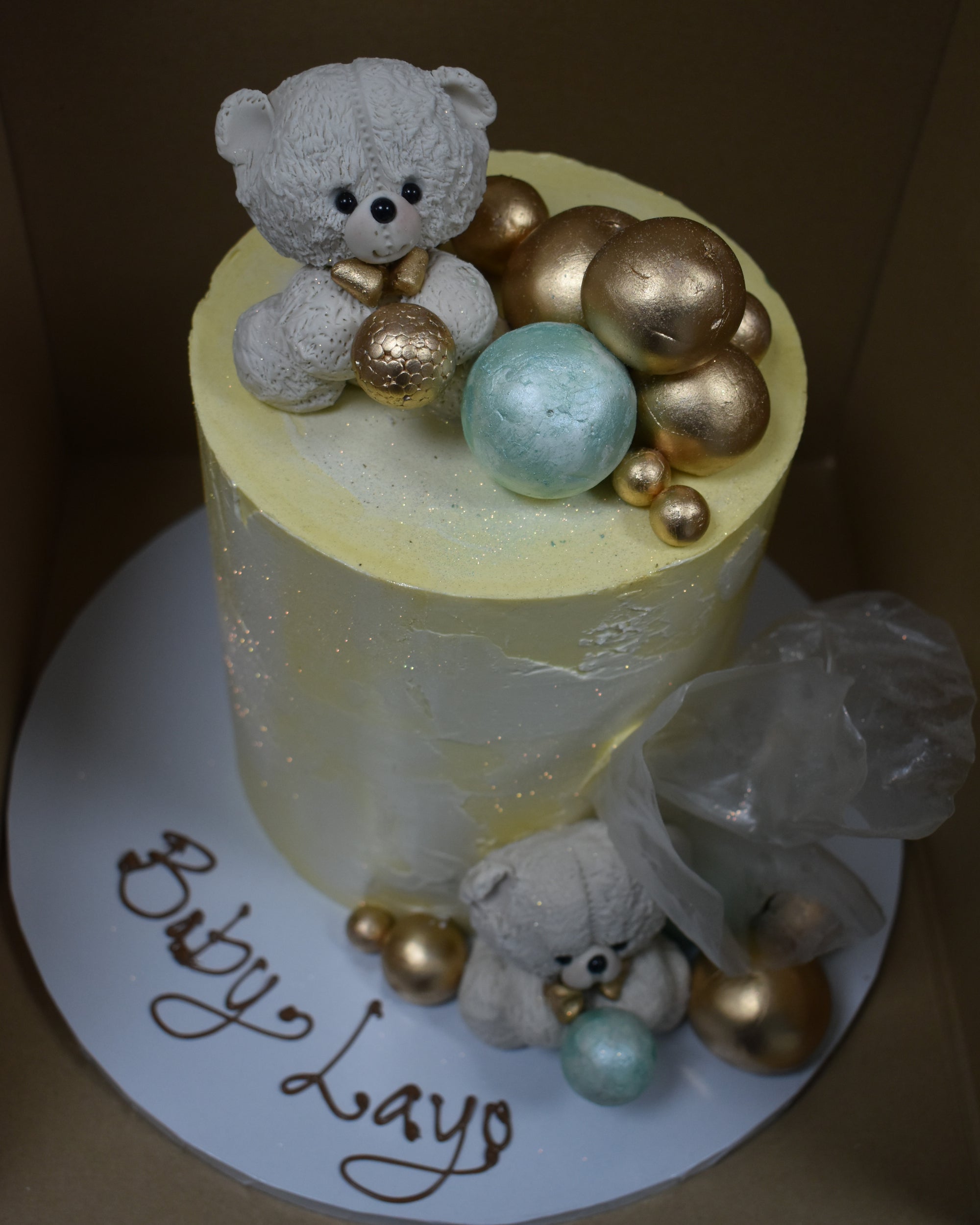 yellow and white warercolour cake with white teddies on top and in front of the cake with a selection of pistachio mint and gold balls at the bottom and top of the cake with Baby Layo written on the cake board with chocolate