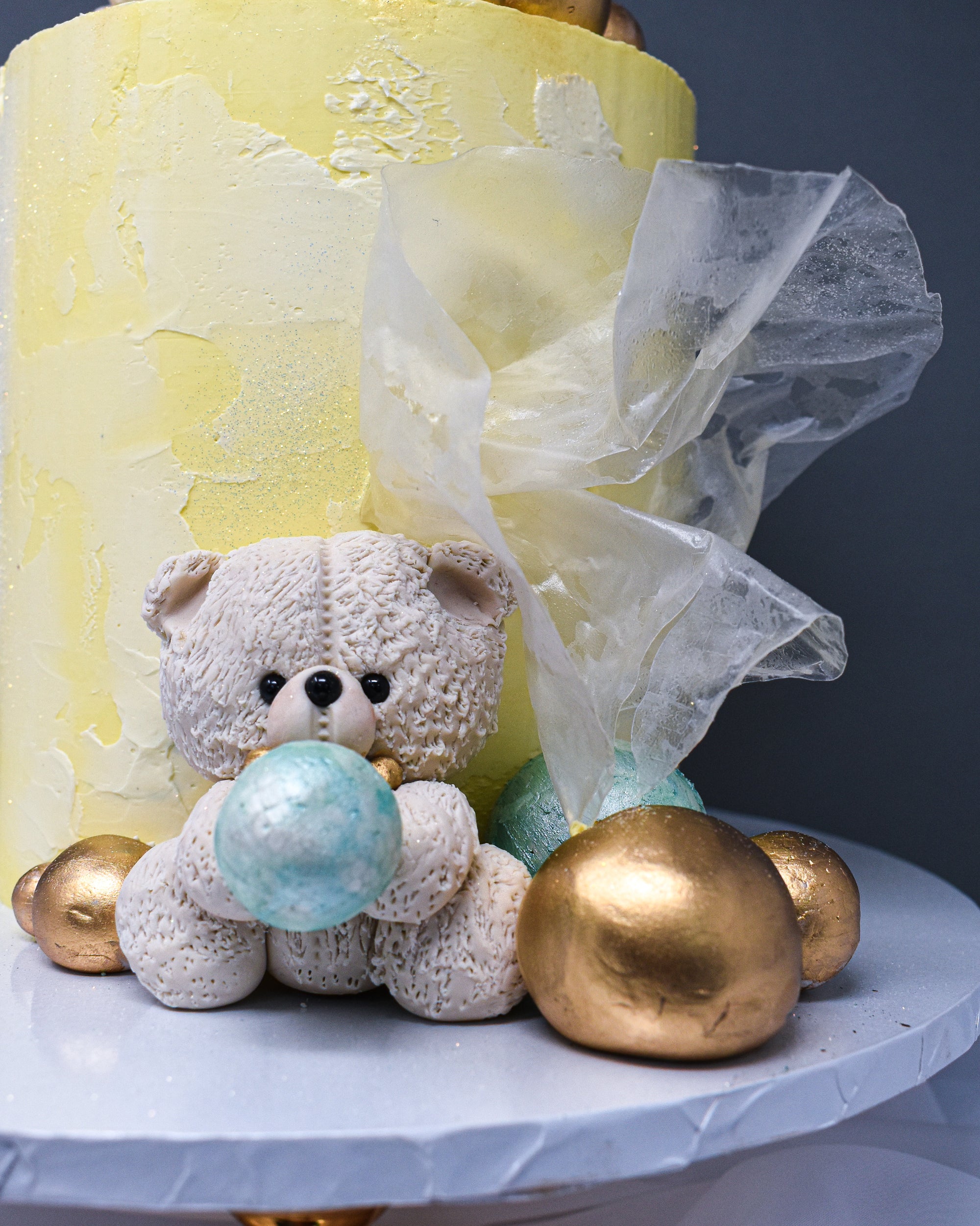 front view of a yellow and white watercolour cake showing a white sugar paste teddy with a gold bow tie, holding a pistachio mint ball surrounded by mixed sizes of gold and pistachio mint balls and a transluscent rice paper sail.