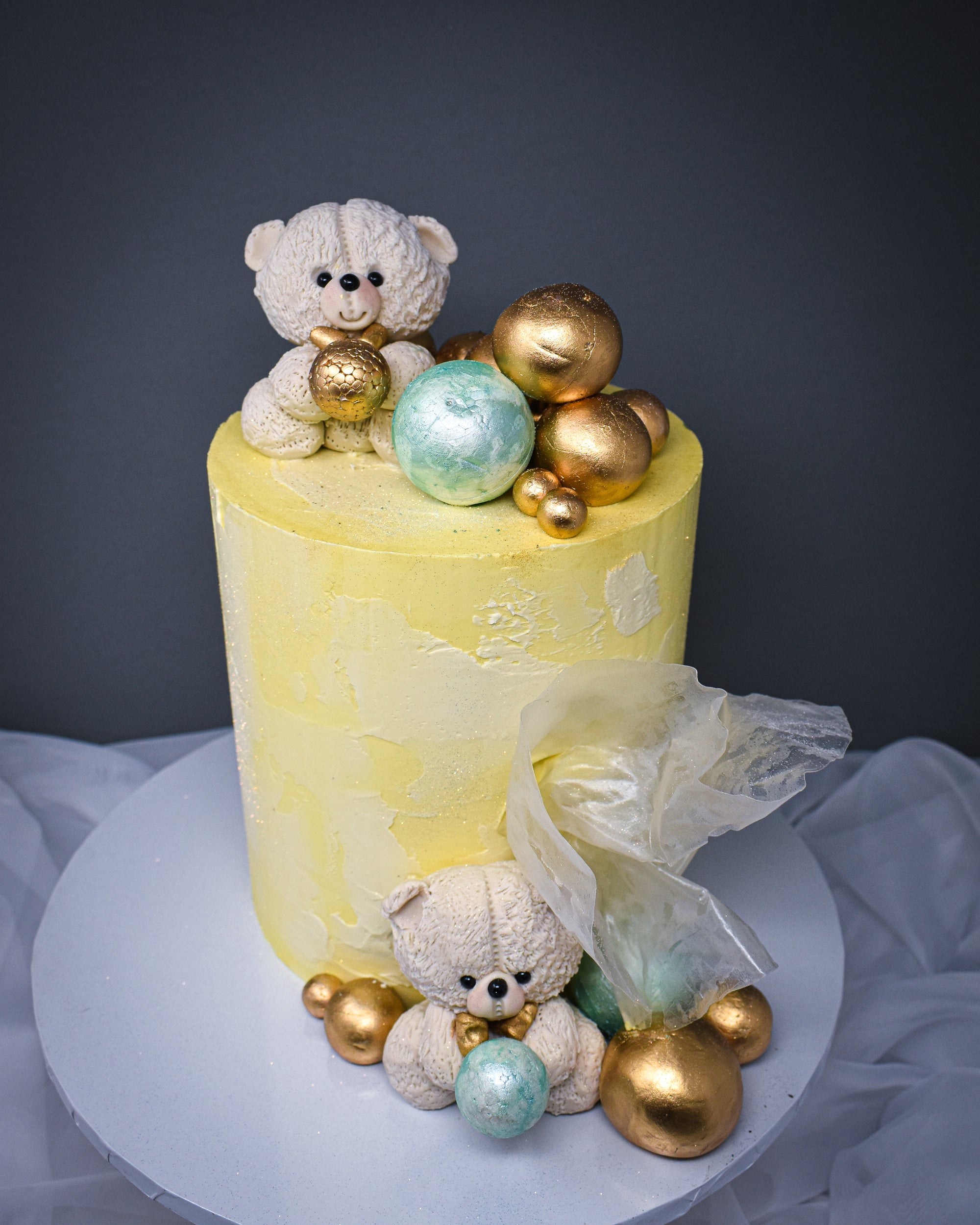 ariel view of yellow and white warercolour cake with white teddies on top and in front of the cake with a selection of pistachio mint and gold balls at the bottom and top of the cake