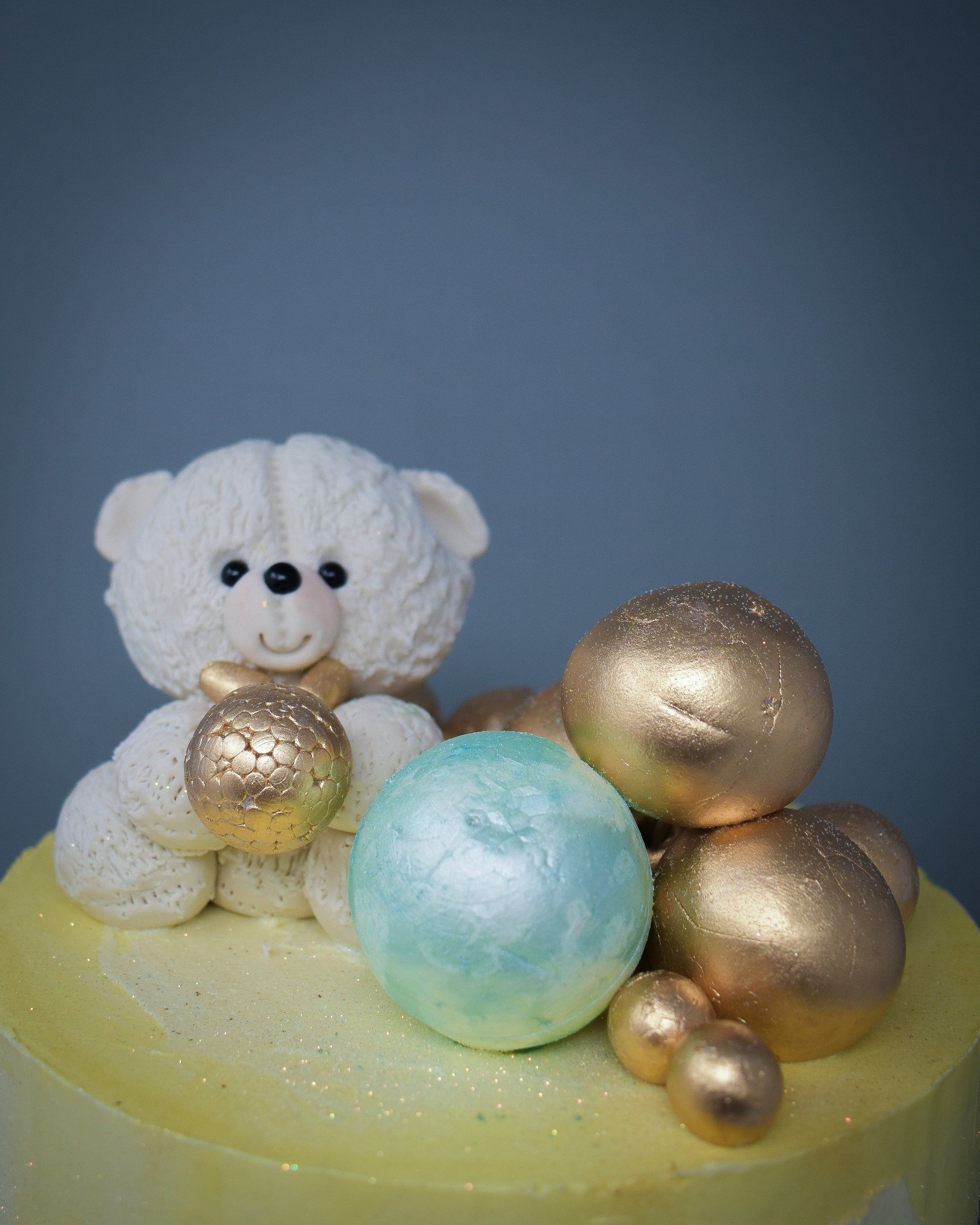 top view of a yellow and white watercolour cake showing a white sugar paste teddy with a gold bow tie, holding a gold ball surrounded by mixed sizes of gold and pistachio mint balls