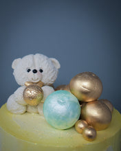 Load image into Gallery viewer, top view of a yellow and white watercolour cake showing a white sugar paste teddy with a gold bow tie, holding a gold ball surrounded by mixed sizes of gold and pistachio mint balls
