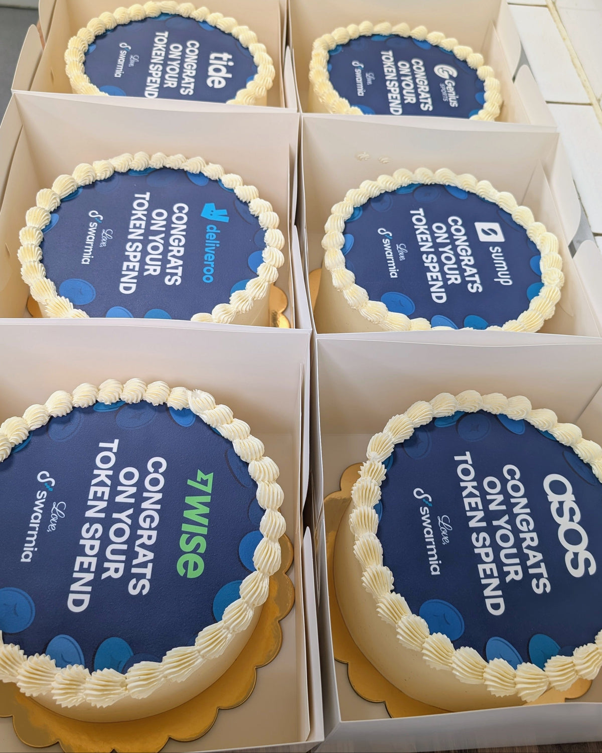 Six round logo cakes decorated with blue and white edible images arranged in two rows inside white cardboard gift boxes.