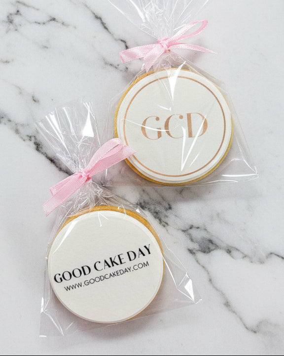 Two round cookies with 'GCD' and 'Good Cake Day' branding in clear packaging with pink ribbons on a light gray background.