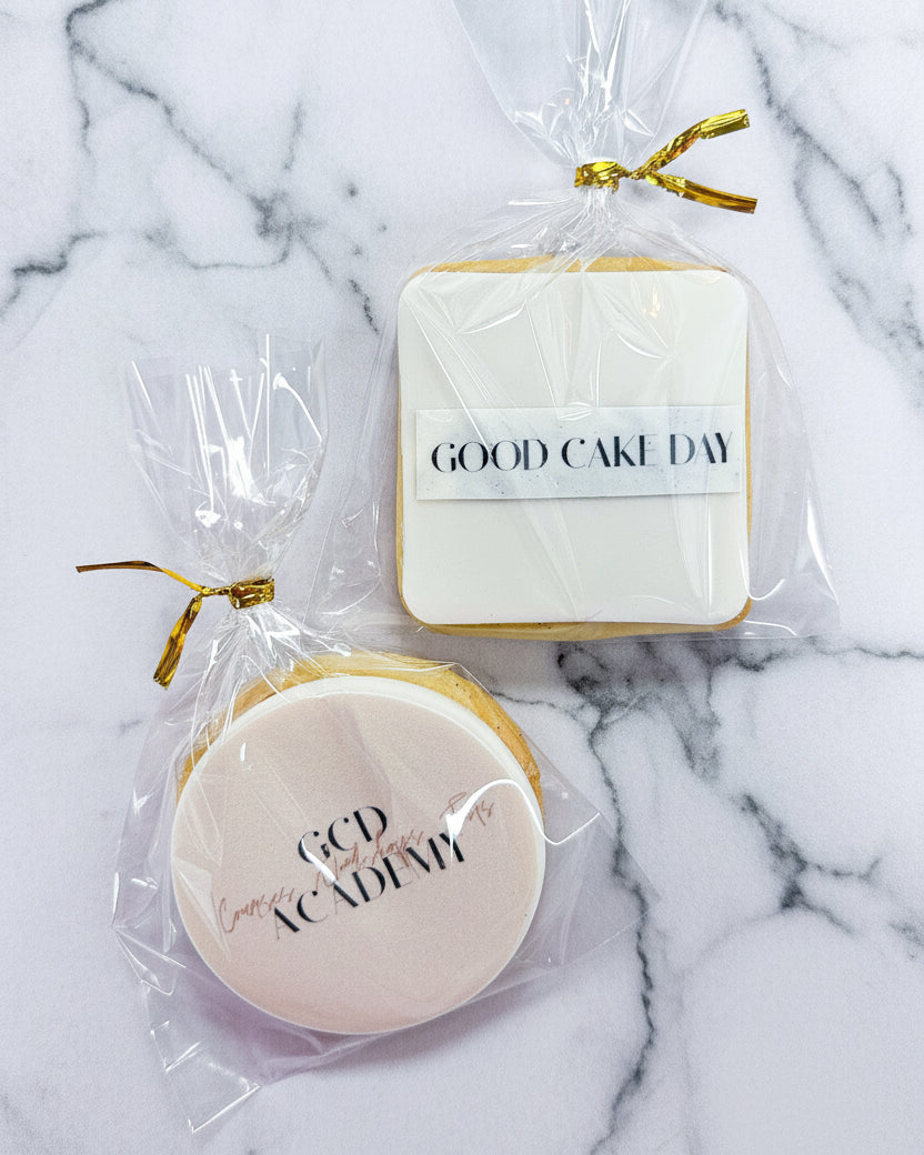 Two cookies with 'GOOD CAKE DAY' text in clear packaging on a marble background. 