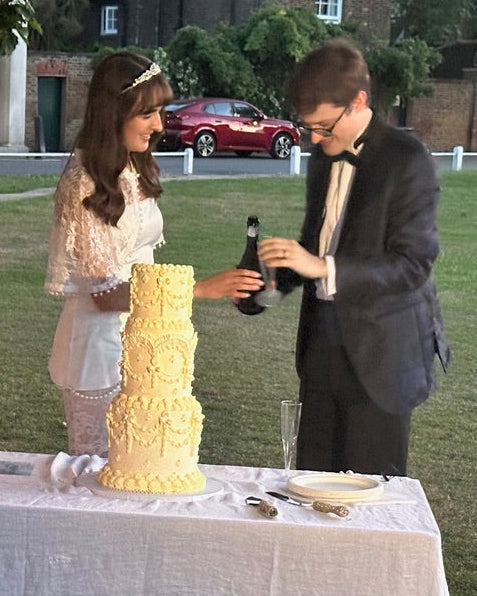 A bride and groom standing outdoors on a grassy lawn beside a table with a white tablecloth, a 3 tiered lambeth cake, a lit candle, and flowers. The groom holds a bottle while the bride holds a glass.