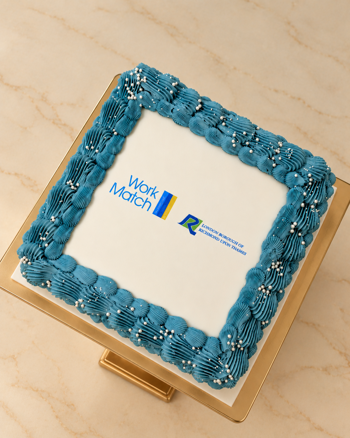 A square white cake with a piped blue border and small white decorative beads, featuring the Work Match logo and Brazilian flag graphic on the top, displayed on a gold cake stand against a beige marble background.
