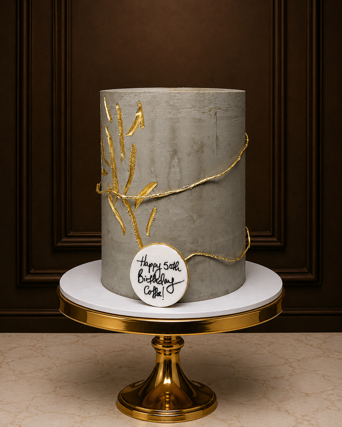 A tall, cylindrical gray cake with textured detailing and gold leaf branch accents, displayed on a white and gold pedestal stand against a dark brown paneled background. A round white topper reads 'Happy 60th Birthday Coffee!'.