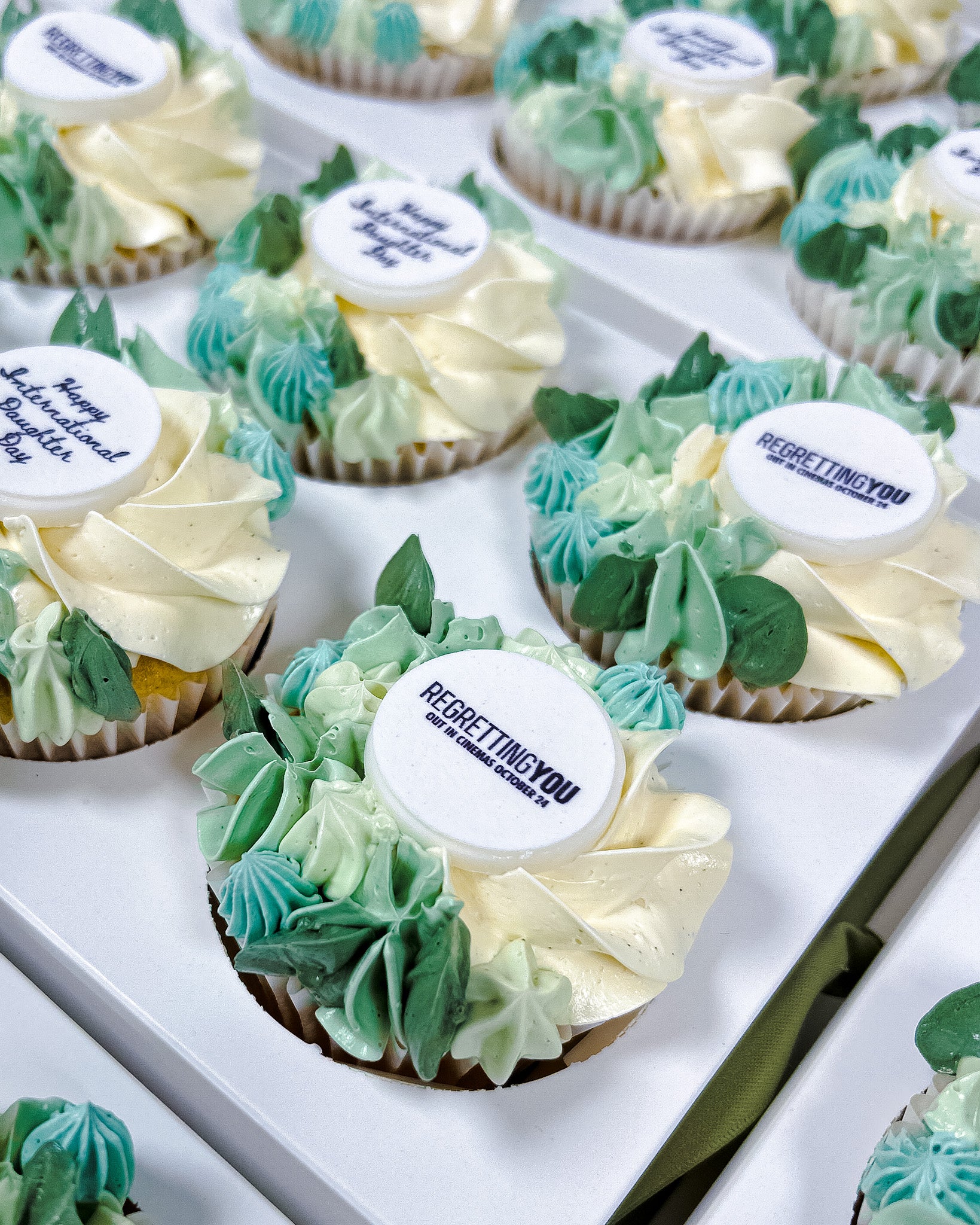 Floral Cupcakes with green and white frosting and 'REGRETTINGYOU' branding, arranged on a white tray.