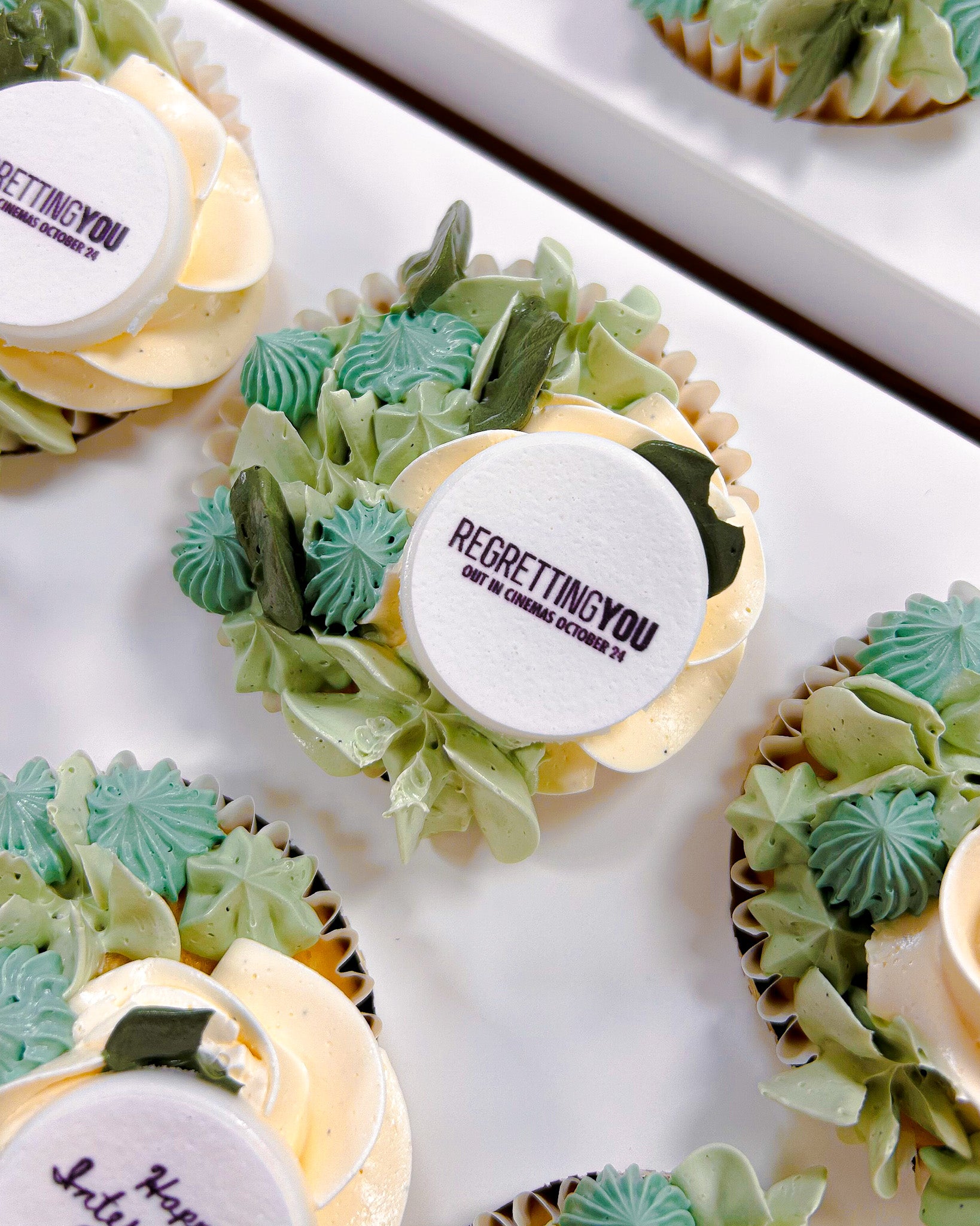 Floral Cupcakes with green and yellow frosting and 'REGRETTING YOU' logo, arranged on a white surface.