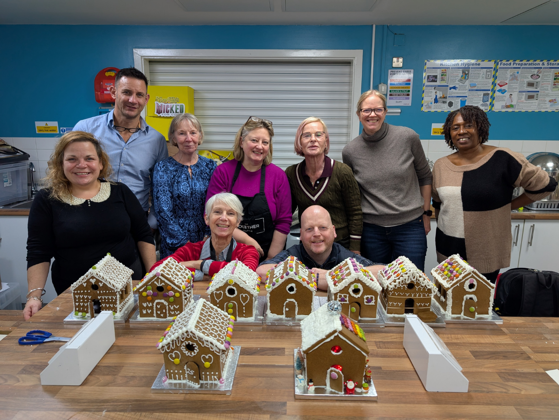 A group of nine people smiling behind a table displaying ten decorated gingerbread houses with white icing roofs and colorful candy details, set in a kitchen environment.