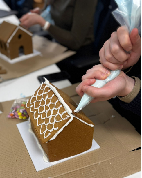 A person piping white icing onto a gingerbread house roof at a table, with candy wrappers and a phone visible in a real workshop or party setting.