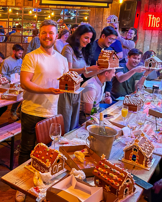 A group of people in a restaurant setting holding decorated gingerbread houses. The scene is warmly lit with ambient lighting, showing a festive atmosphere with tables covered in food and drinks.