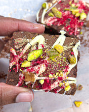 Load image into Gallery viewer, Picture of a hand holding up Dark chocolate brownies infused with rose water &amp; white chocolate pieces adorned with white chocolate, crushed pistachios and freeze dried raspberries Media  
