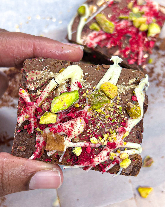 Picture of a hand holding up Dark chocolate brownies infused with rose water & white chocolate pieces adorned with white chocolate, crushed pistachios and freeze dried raspberries Media  