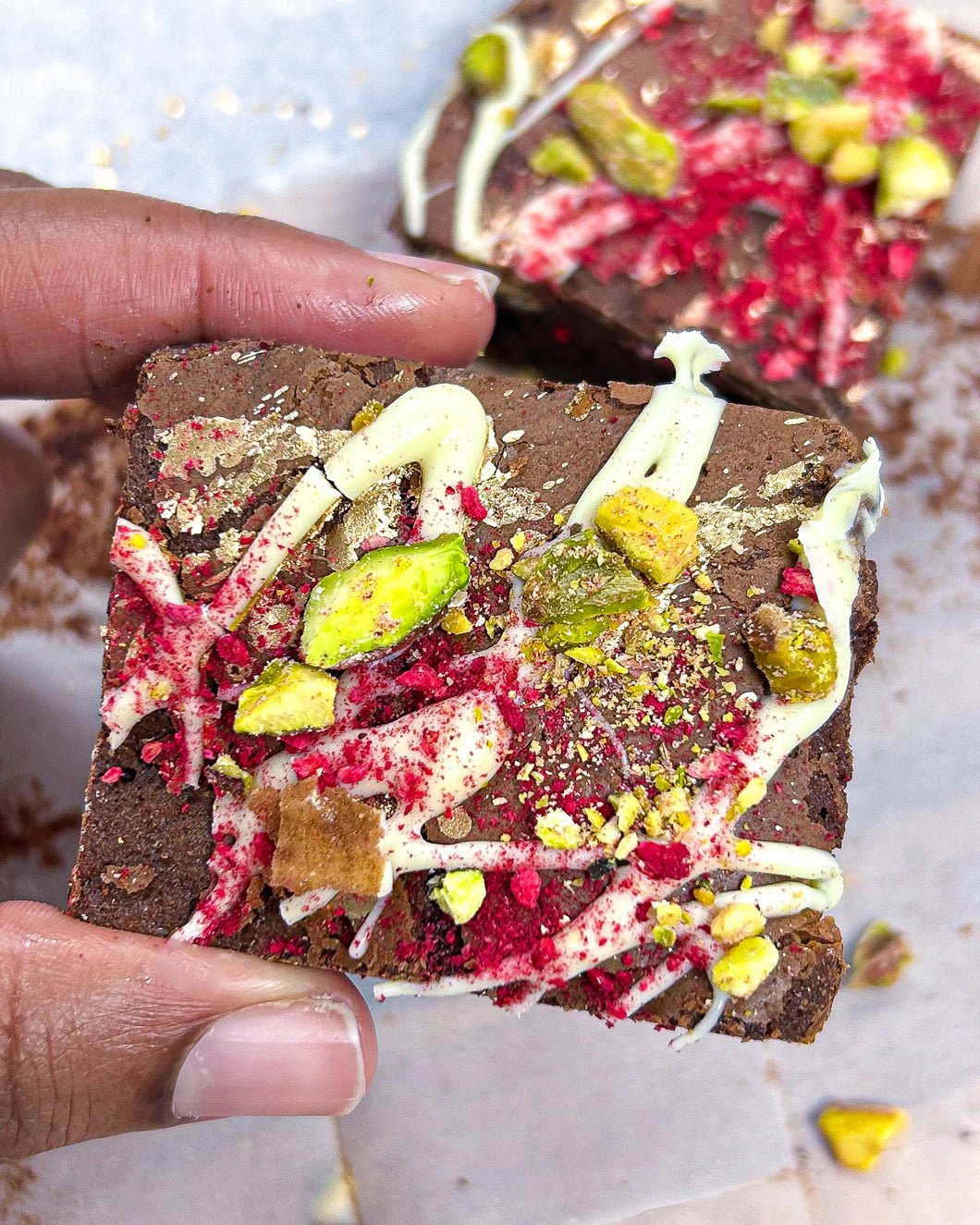 Picture of a hand holding up Dark chocolate brownies infused with rose water & white chocolate pieces adorned with white chocolate, crushed pistachios and freeze dried raspberries Media  