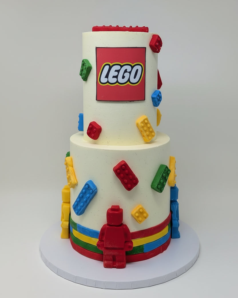 Two-tiered cake decorated with colorful LEGO bricks and a LEGO logo on a white background.
