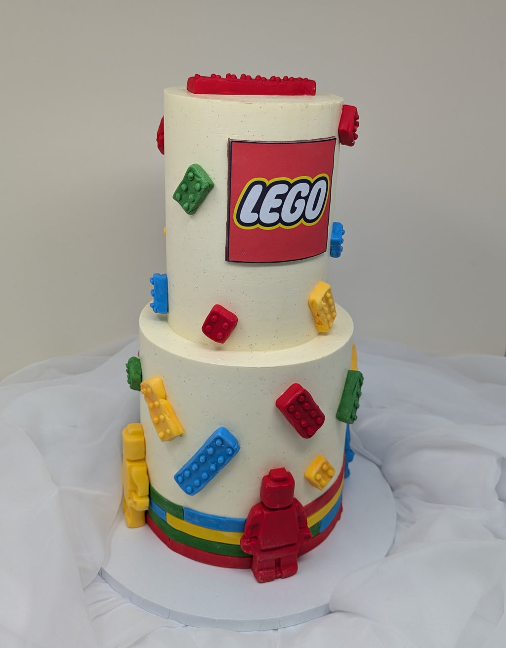 Two-tiered cake decorated with colorful LEGO bricks and a large LEGO logo on a white background.