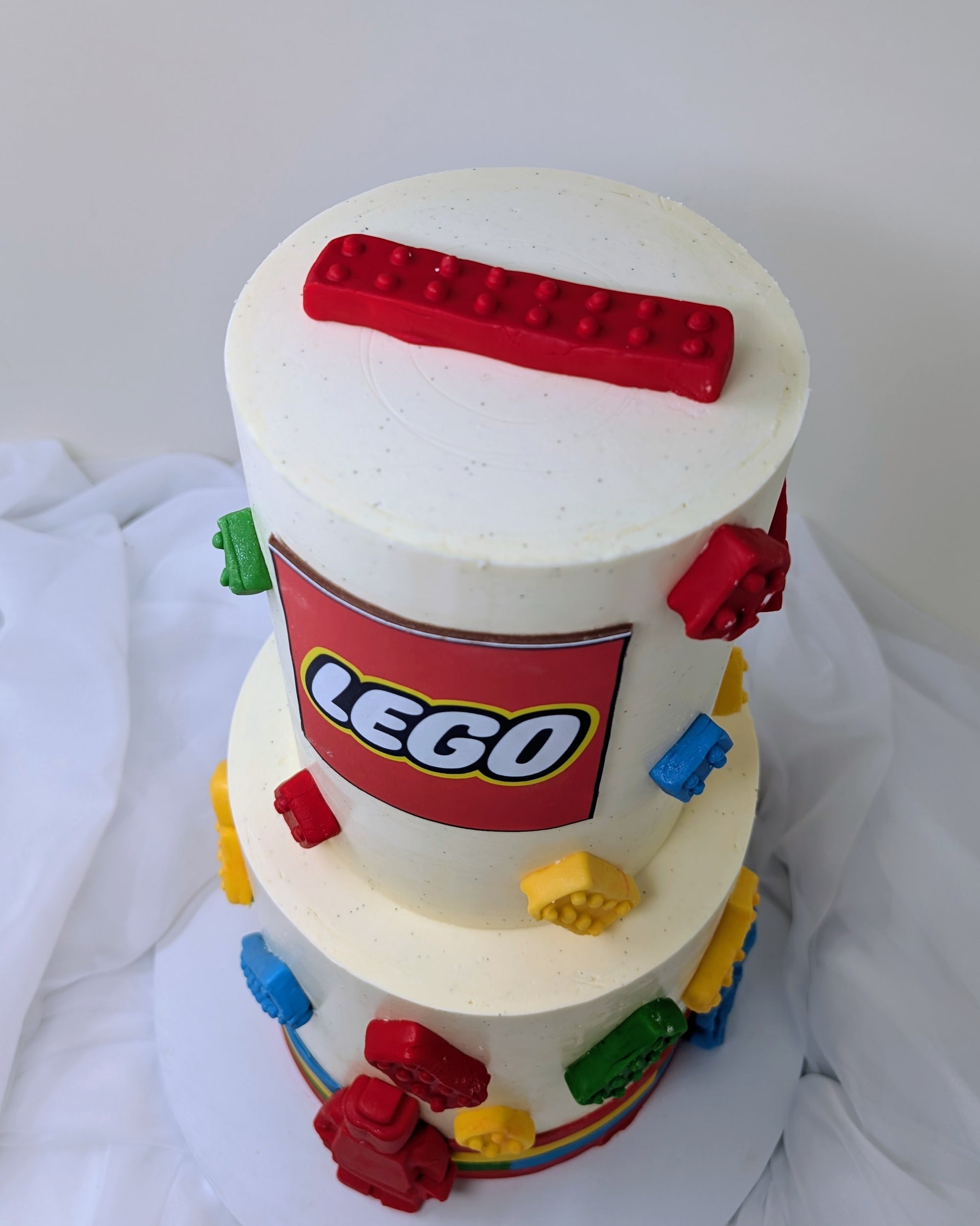Two-tiered cake decorated with LEGO bricks and a prominent LEGO logo on a white background.