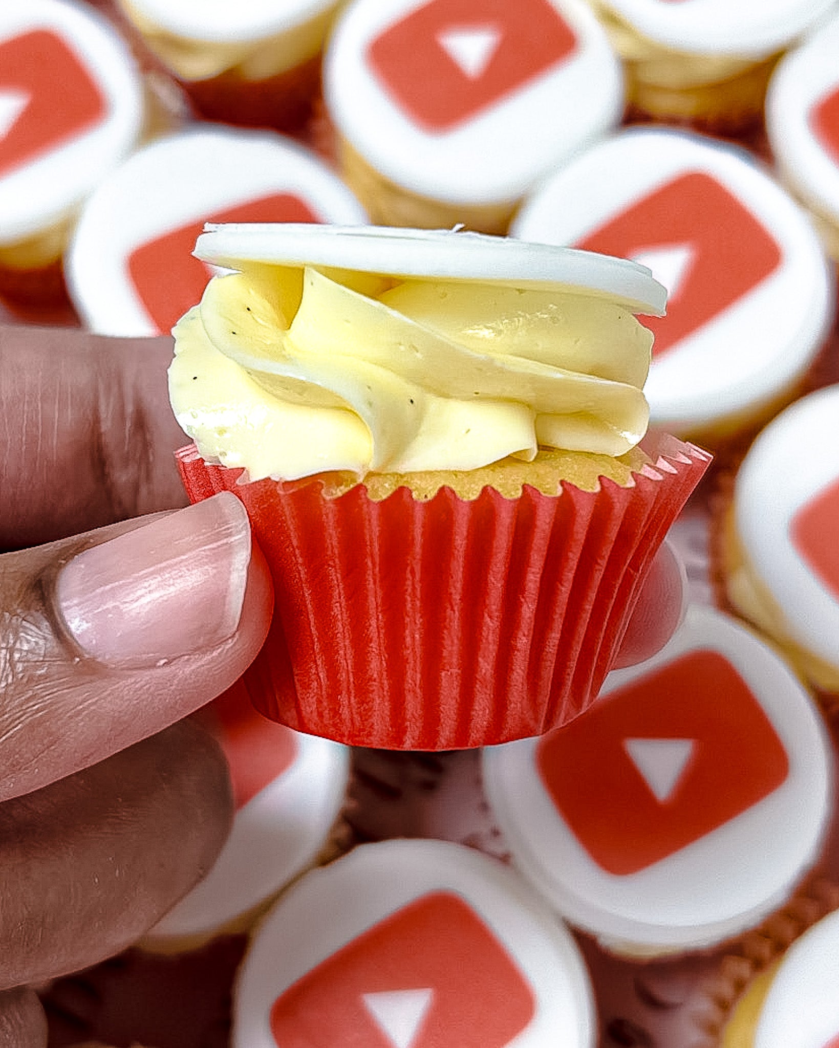 Close-up of a mini cupcake with cream frosting and YouTube logo, and red wrapper held by a hand, surrounded by cupcakes with similar designs.