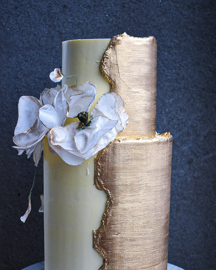 A two-tiered cake with a split design: one side is smooth cream-colored frosting, while the other side is wrapped in textured gold leaf. Large white artificial flowers are attached to the left side, and the cake sits on a white marble round stand against a dark textured background.