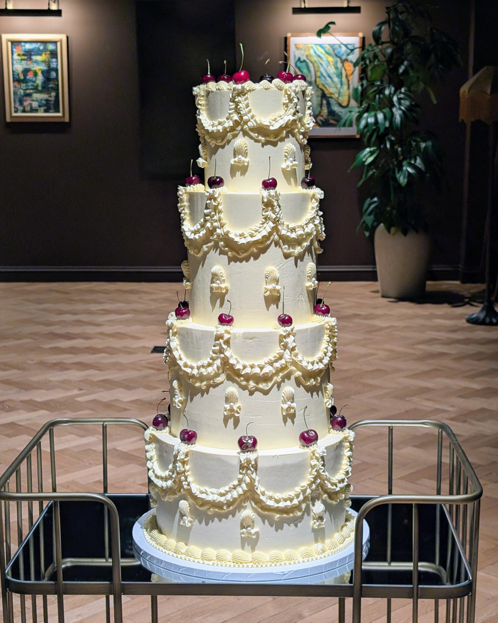 A four-tier white wedding cake decorated with intricate cream buttercream patterns and small red cherry accents, displayed on a metal cart in an indoor event space.
