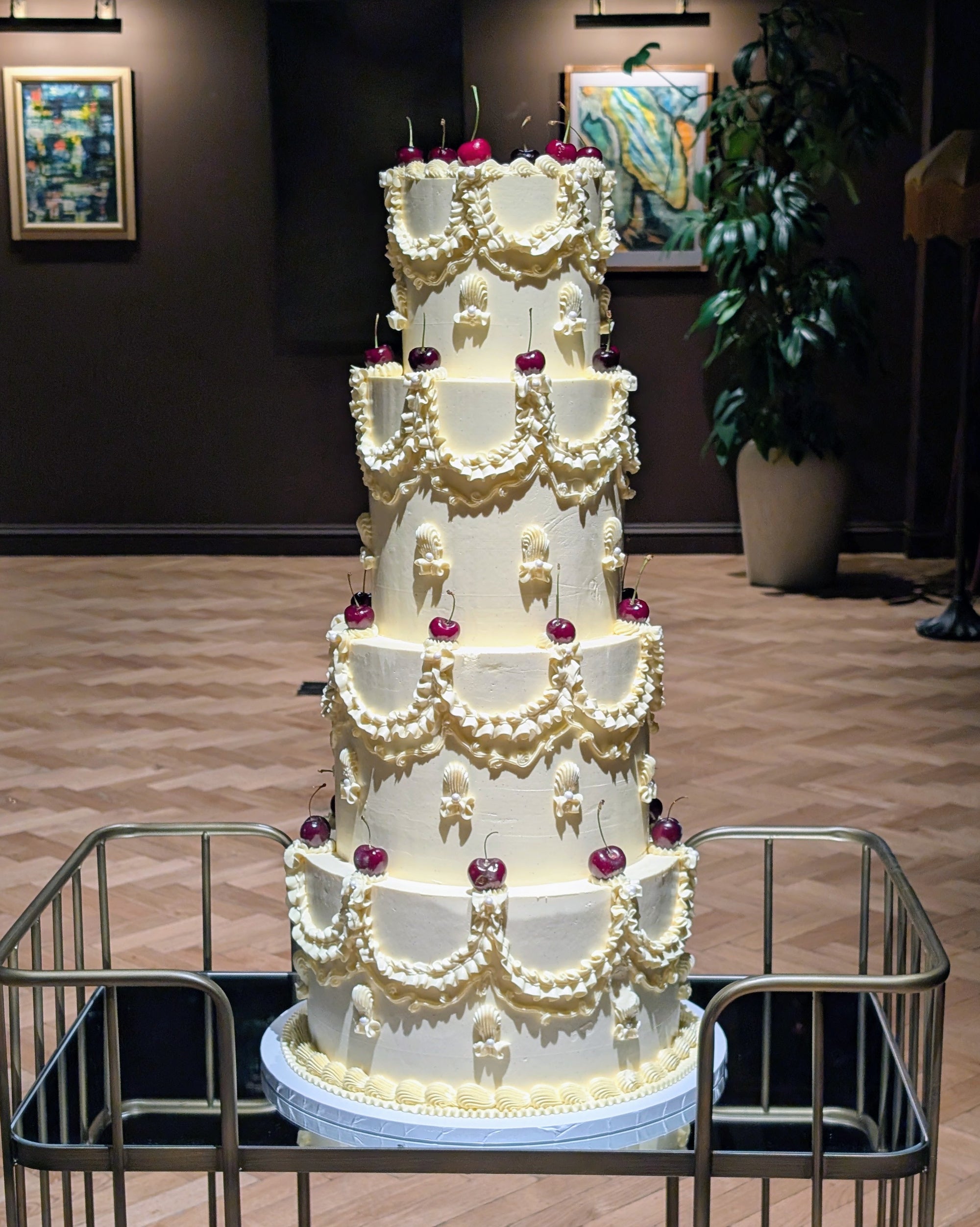 A four-tier white wedding cake decorated with intricate cream buttercream patterns and small red cherry accents, displayed on a metal cart in an indoor event space.