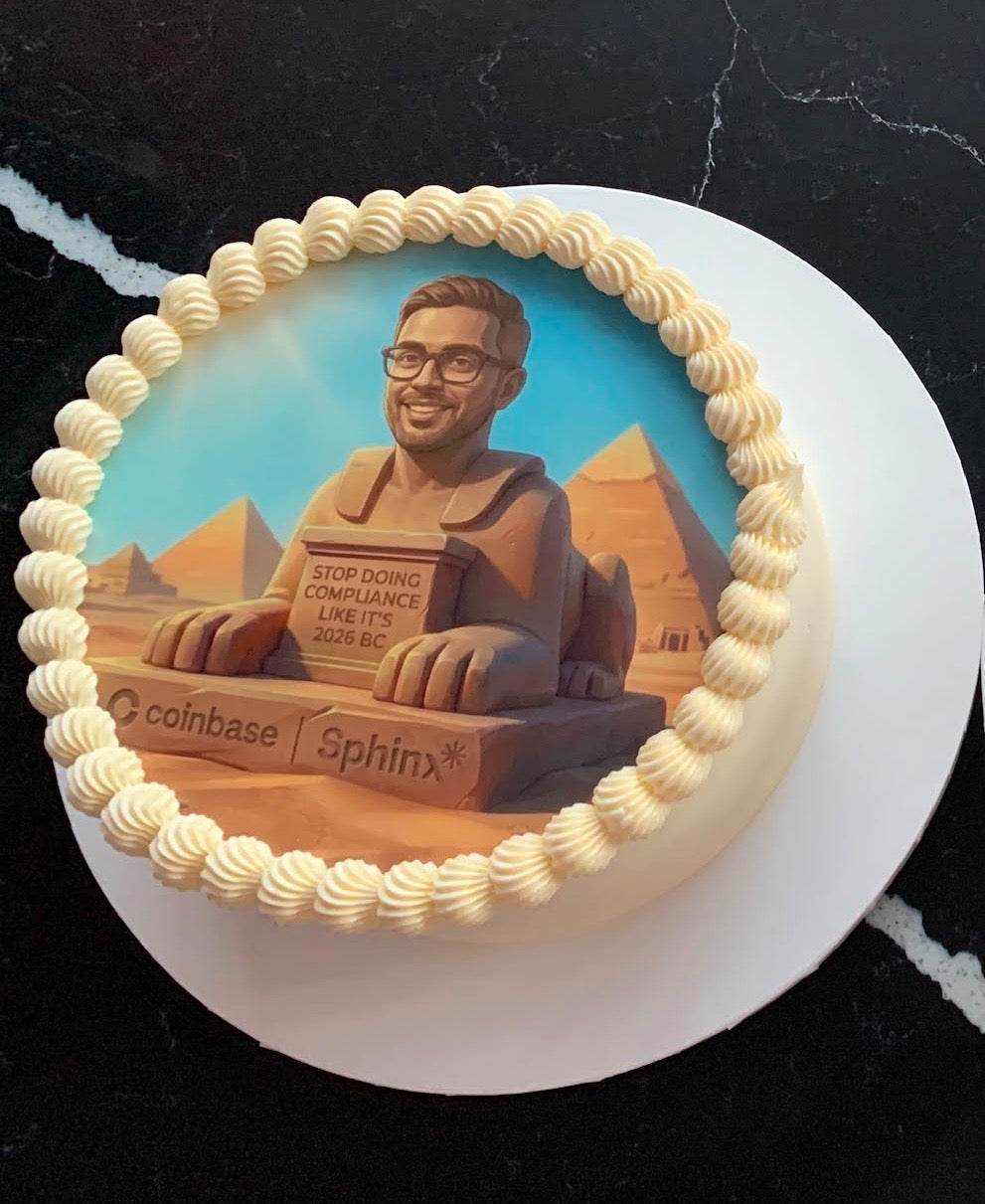 A round cake decorated with piped cream frosting around the edge and a printed or sculpted illustration of a sphinx statue in an Egyptian desert scene, with the words 'coinbase' and 'Sphinx' visible on the front, set on a white plate against a dark marble surface.