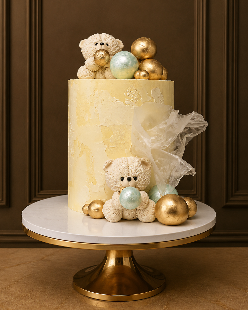 A custom cylindrical cake with textured cream frosting, decorated with two small cream teddy bears holding gold and light blue ornaments, gold metallic balls, and a piece of sheer tulle, displayed on a round white and gold cake stand against a dark brown paneled wall.