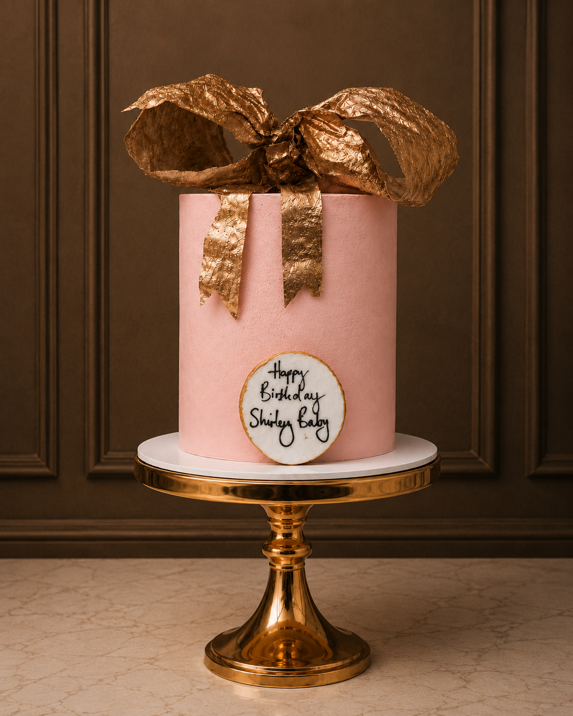 A pink frosted birthday cake shaped like a gift box with a large metallic gold bow on top, displayed on a gold pedestal stand against a dark brown paneled wall.