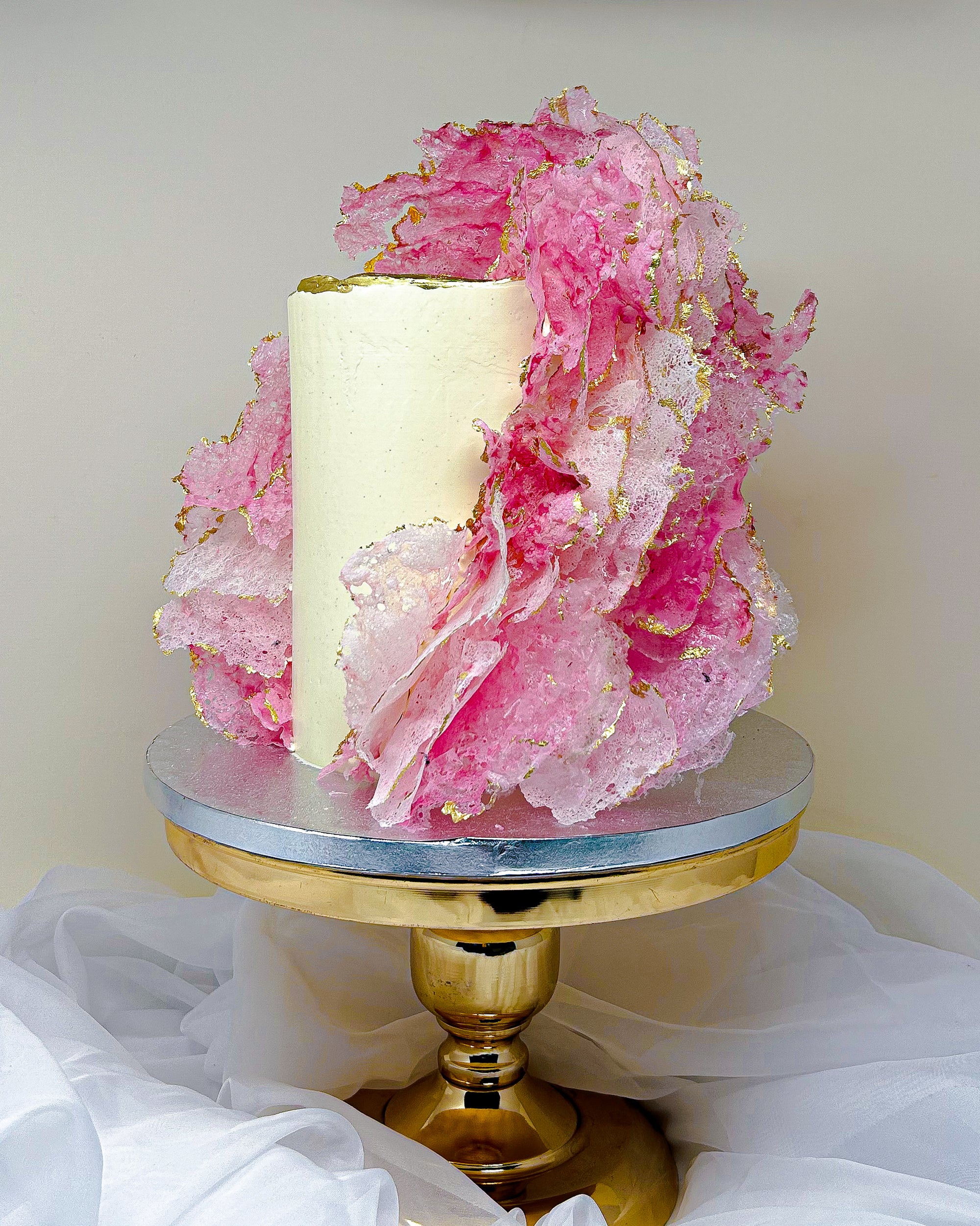 Decorative cake with pink floral elements on a gold stand against a white background