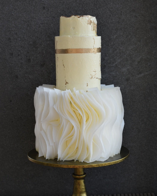A three-tier wedding cake with a cream-colored top tier and a white ruffled bottom tier, decorated with gold leaf and a gold band, displayed on a gold cake stand against a dark grey background.