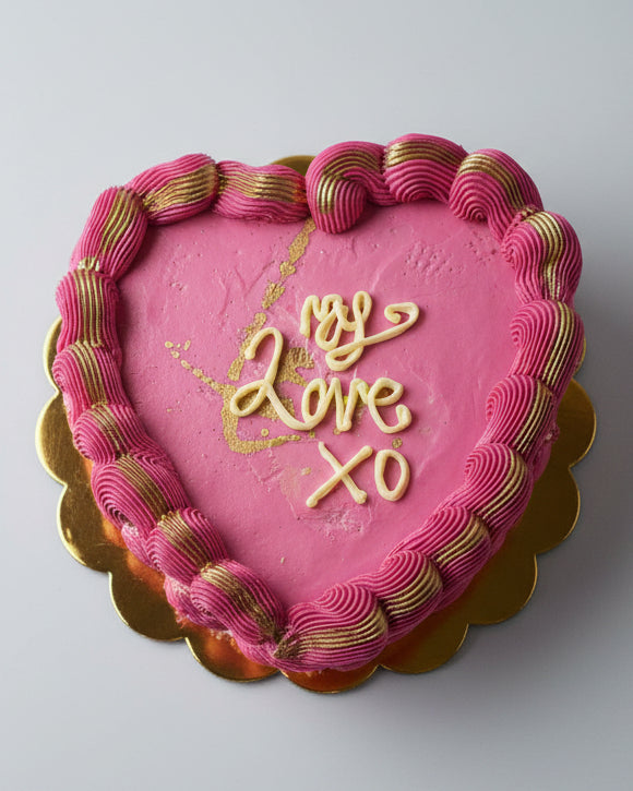 A heart-shaped cake with pink buttercream frosting and the words 'My Love XO' written on top in decorative script, surrounded by gold buttercream designs.