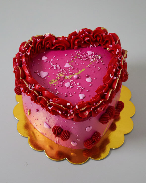 A heart-shaped cake decorated with pink and red buttercream and heart sprinkles, presented on a yellow platter.