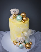 Load image into Gallery viewer, ariel view of yellow and white warercolour cake with white teddies on top and in front of the cake with a selection of pistachio mint and gold balls at the bottom and top of the cake
